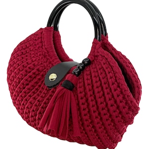 May include: A red crocheted handbag with black handles and a tassel. The bag features a black leather accent with a gold-colored button. The handles are glossy and round. The bag's body is a solid, textured red.