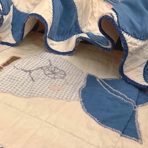 May include: A quilted fabric piece featuring white and blue panels joined with dark blue zigzag stitching. The surface displays a light blue and white gingham patch with a gray line-drawn illustration of a figure and a small pink accent.