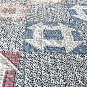 May include: A patchwork quilt with a repeating pattern of geometric shapes. The quilt features squares with house-like designs in red, white, and blue, set against a background of small blue floral prints. The quilt is made of fabric.