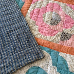 May include: A quilted textile featuring a patchwork design with a large pink floral motif, blue and white plaid sections, and solid orange borders. The fabric displays intricate stitching patterns throughout the surface, combining various geometric shapes and floral patterns in shades of pink, blue, white, and orange.