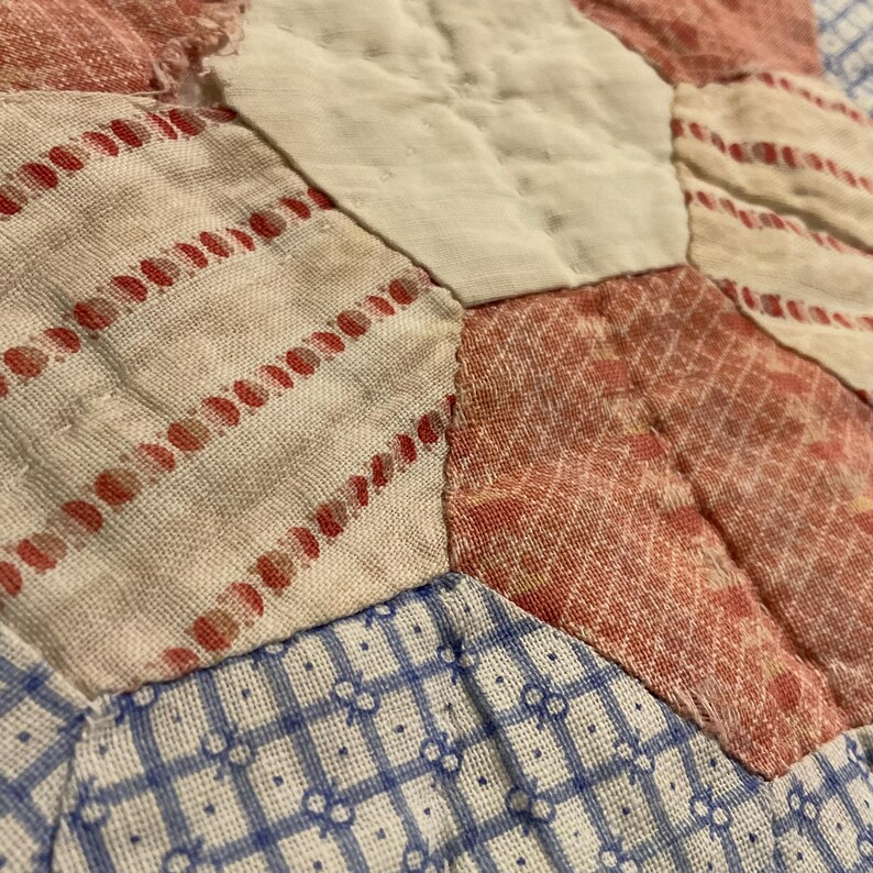 K&ouml;nnte beinhalten: Ein Patchwork-Quilt mit einem hexagonalen Muster aus verschiedenen Stoffst&uuml;cken. Das Design umfasst cremewei&szlig;e Stoffe, rot-wei&szlig; gestreifte Stoffe, rot gemusterte Stoffe sowie blau-wei&szlig; karierte Stoffe, die miteinander vern&auml;ht sind.