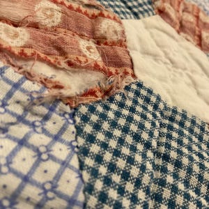 K&ouml;nnte beinhalten: Ein Patchwork-Quilt mit verschiedenen Stoffquadraten in blau-wei&szlig;em Vichy-Karo, blau-wei&szlig;en Blumenmustern und rot-wei&szlig; gemustertem Stoff. Das Textil weist sichtbare Gebrauchsspuren mit ausgefransten Kanten und losen F&auml;den auf.