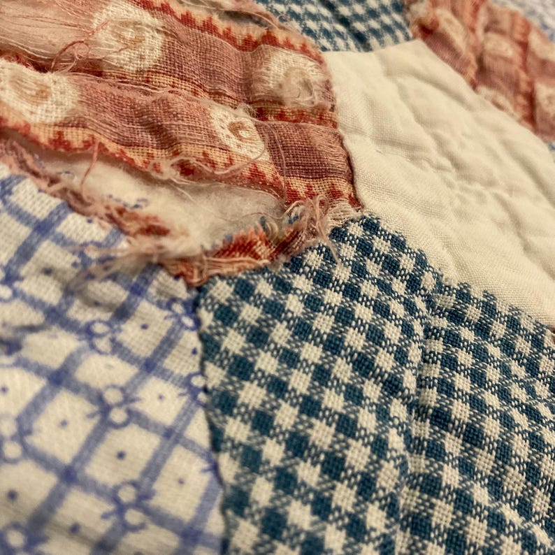 K&ouml;nnte beinhalten: Ein Patchwork-Quilt mit verschiedenen Stoffquadraten in blau-wei&szlig;em Vichy-Karo, blau-wei&szlig;en Blumenmustern und rot-wei&szlig; gemustertem Stoff. Das Textil weist sichtbare Gebrauchsspuren mit ausgefransten Kanten und losen F&auml;den auf.