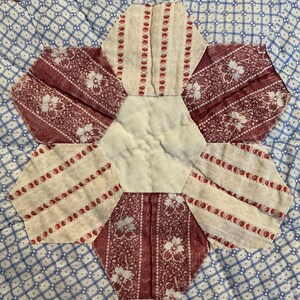 K&ouml;nnte beinhalten: Ein Quiltblock mit einem zentralen cremefarbenen Sechseck, umgeben von sechs abwechselnden Sechsecken. Drei Sechsecke zeigen ein dunkelrotes Blumenmuster, w&auml;hrend die anderen drei einen cremefarbenen Stoff mit vertikalen roten Punktstreifen aufweisen.