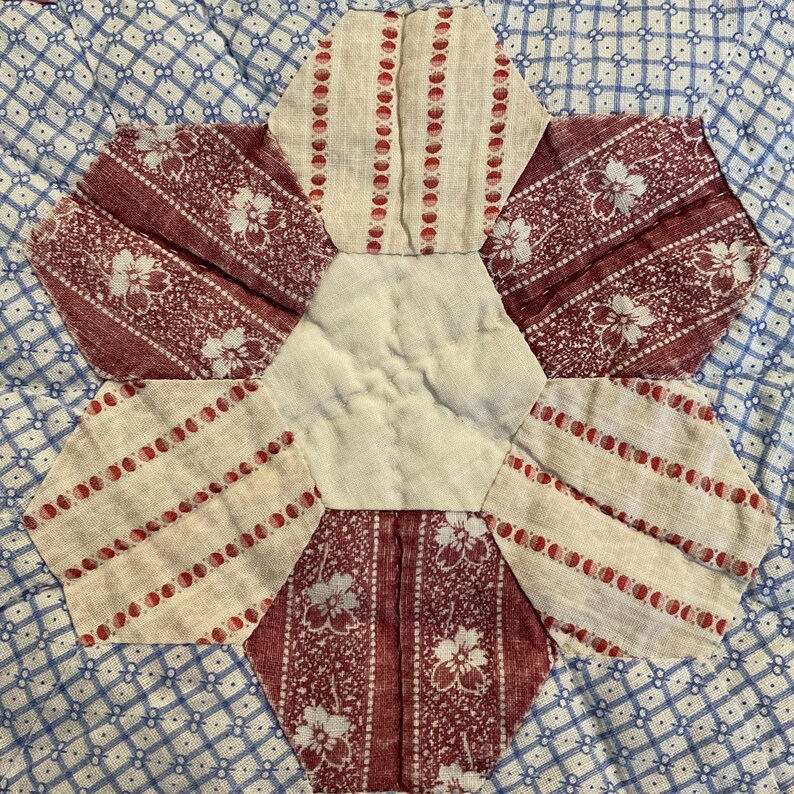K&ouml;nnte beinhalten: Ein Quiltblock mit einem zentralen cremefarbenen Sechseck, umgeben von sechs abwechselnden Sechsecken. Drei Sechsecke zeigen ein dunkelrotes Blumenmuster, w&auml;hrend die anderen drei einen cremefarbenen Stoff mit vertikalen roten Punktstreifen aufweisen.