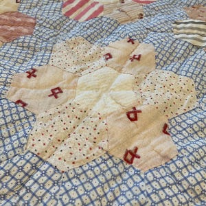 K&ouml;nnte beinhalten: Ein Patchwork-Quilt mit einem hexagonalen Muster und einem zentralen cremefarbenen Blumenmotiv. Die Stoffst&uuml;cke zeigen verschiedene Drucke, darunter blau-wei&szlig;e Gitter, rote und schwarze Punkte sowie Streifen, akzentuiert durch rote Handstickereien.
