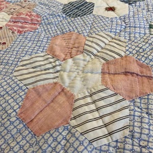 K&ouml;nnte beinhalten: Ein Patchwork-Quilt mit einem hexagonalen Blumenmuster aus verschiedenen Stoffresten. Das Design besteht aus abwechselnden rosa, wei&szlig;en und schwarz gestreiften Stoffsechsecken, die ein zentrales wei&szlig;es Sechseck umgeben, alles auf einem blau-wei&szlig; gemusterten Hintergrund.