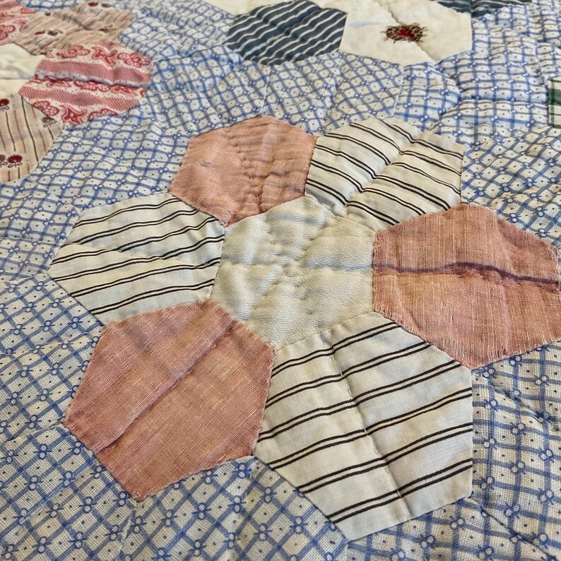 K&ouml;nnte beinhalten: Ein Patchwork-Quilt mit einem hexagonalen Blumenmuster aus verschiedenen Stoffresten. Das Design besteht aus abwechselnden rosa, wei&szlig;en und schwarz gestreiften Stoffsechsecken, die ein zentrales wei&szlig;es Sechseck umgeben, alles auf einem blau-wei&szlig; gemusterten Hintergrund.