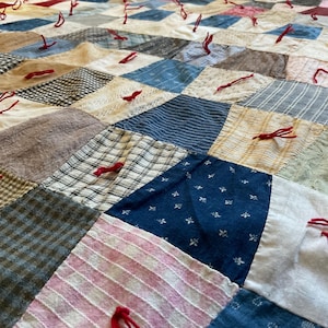 May include: A handmade quilt with a patchwork design. The quilt features various fabric squares in shades of blue, beige, pink, and gray, stitched together. Red thread accents are visible throughout the quilt, adding a pop of color and texture.