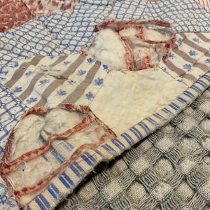 K&ouml;nnte beinhalten: Ein Patchwork-Quilt mit verschiedenen Stoffmustern in Blau, Wei&szlig; und Rot. Das Textil weist deutliche Abnutzungserscheinungen auf, mit ausgefransten Kanten und wei&szlig;er Baumwollf&uuml;llung, die durch Risse in den Stoffquadraten sichtbar ist.