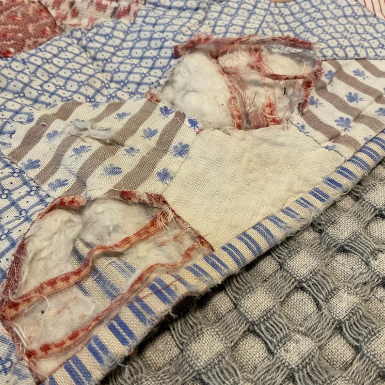 K&ouml;nnte beinhalten: Ein Patchwork-Quilt mit verschiedenen Stoffmustern in Blau, Wei&szlig; und Rot. Das Textil weist deutliche Abnutzungserscheinungen auf, mit ausgefransten Kanten und wei&szlig;er Baumwollf&uuml;llung, die durch Risse in den Stoffquadraten sichtbar ist.