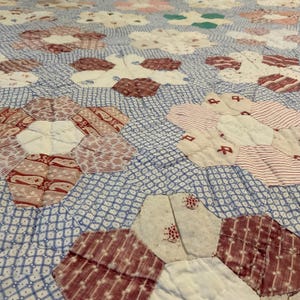K&ouml;nnte beinhalten: Ein Steppdecke mit einem sich wiederholenden Muster aus sechseckigen Blumenmotiven. Die Blumen bestehen aus verschiedenen roten, wei&szlig;en und cremefarbenen Stoffst&uuml;cken mit diversen Drucken auf einem Hintergrund aus blau-wei&szlig; kariertem Stoff.