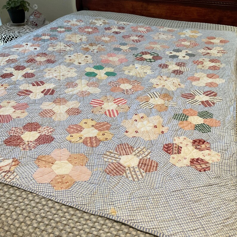 K&ouml;nnte beinhalten: Eine Patchwork-Decke mit einem sich wiederholenden Muster aus sechseckigen Blumenmotiven in verschiedenen Rot-, Rosa-, Orange-, Gr&uuml;n- und Beiget&ouml;nen. Der Hintergrundstoff besteht aus einem hellblauen und wei&szlig;en Karomuster mit sichtbaren N&auml;hten.