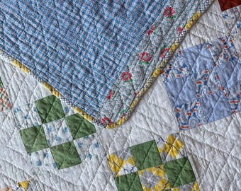 Könnte beinhalten: Ein gestepptes Textil mit einem Patchwork-Design aus weißen, grünen, gelben und blauen Stoffquadraten. Der obere Bereich hat ein blau-weißes Karomuster mit einer Blumenbordüre, während der Rest verschiedene geometrische Muster aufweist.