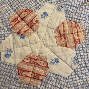K&ouml;nnte beinhalten: Ein gestepptes Textil mit einem zentralen wei&szlig;en Sechseck, das von abwechselnden sechseckigen Flicken umgeben ist. Das Muster umfasst wei&szlig;en Stoff mit blauen Blumenmustern, rot-wei&szlig;en Paisley-Streifen und einem blau-wei&szlig; karierten Hintergrund.