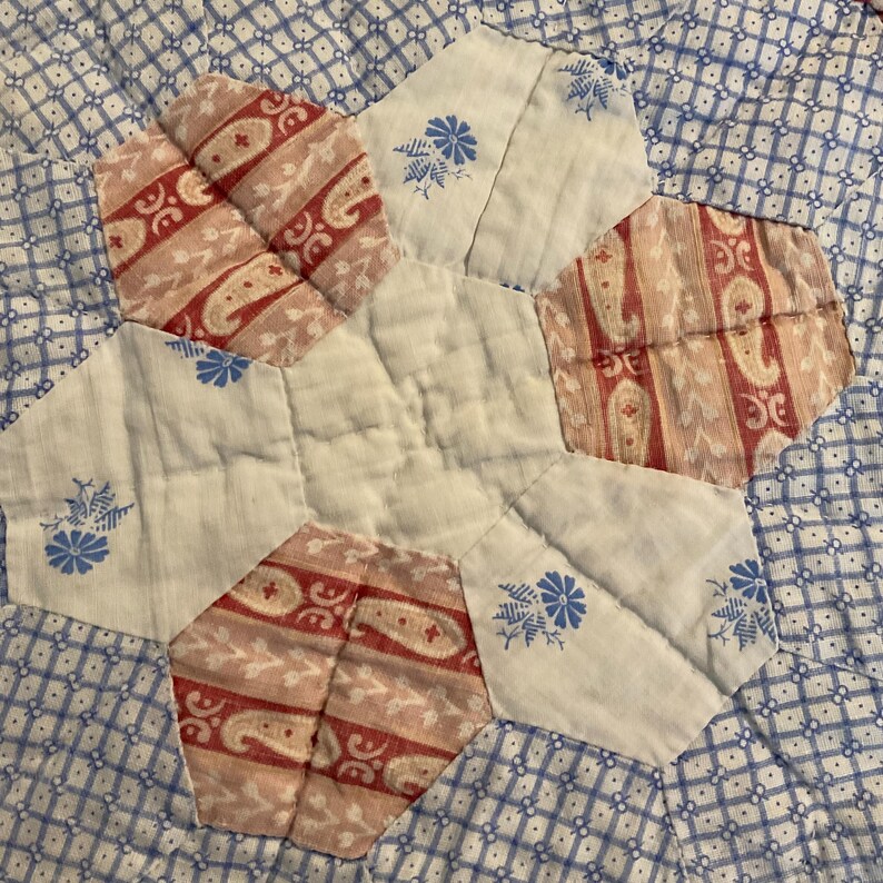 K&ouml;nnte beinhalten: Ein gestepptes Textil mit einem zentralen wei&szlig;en Sechseck, das von abwechselnden sechseckigen Flicken umgeben ist. Das Muster umfasst wei&szlig;en Stoff mit blauen Blumenmustern, rot-wei&szlig;en Paisley-Streifen und einem blau-wei&szlig; karierten Hintergrund.