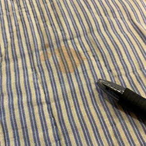 K&ouml;nnte beinhalten: Ein wei&szlig;er Stoff mit d&uuml;nnen blauen vertikalen Streifen, der einen orangefarbenen Fleck aufweist. Ein schwarzer Kugelschreiber mit einer durchsichtigen Kunststoffkappe ist unten rechts auf dem Stoff platziert, um die Gr&ouml;&szlig;e des Flecks zu verdeutlichen.