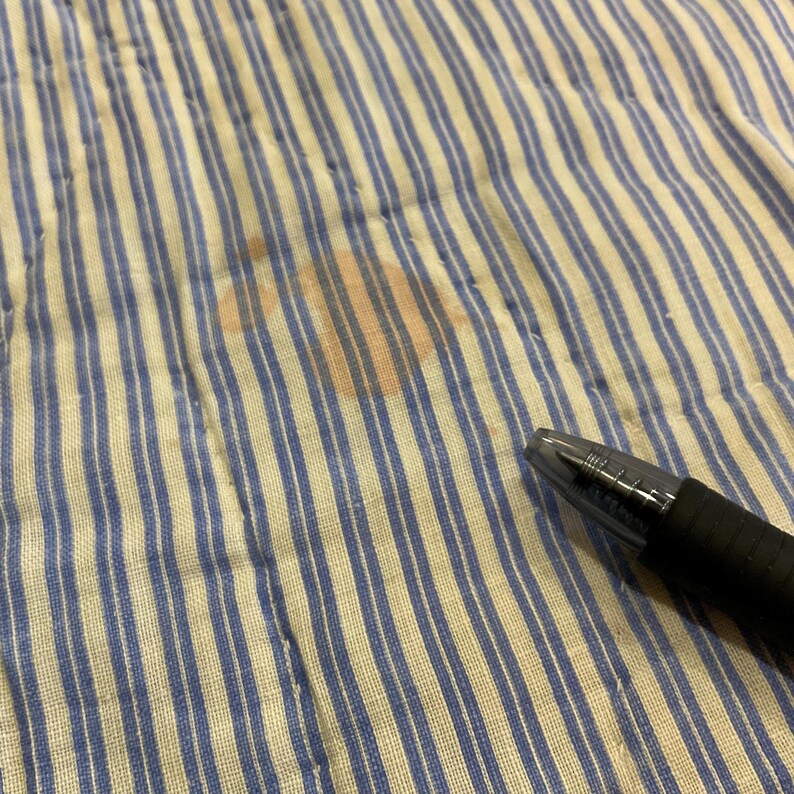 K&ouml;nnte beinhalten: Ein wei&szlig;er Stoff mit d&uuml;nnen blauen vertikalen Streifen, der einen orangefarbenen Fleck aufweist. Ein schwarzer Kugelschreiber mit einer durchsichtigen Kunststoffkappe ist unten rechts auf dem Stoff platziert, um die Gr&ouml;&szlig;e des Flecks zu verdeutlichen.