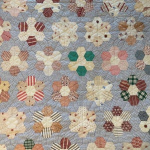 K&ouml;nnte beinhalten: Ein Quilt mit einem sich wiederholenden Muster aus hexagonalen Blumenmotiven. Jede Blume besteht aus sechs Stoffsechsecken in verschiedenen Farben wie Creme, Beige, Rosa, Rot und Gr&uuml;n, auf einem hellblauen und wei&szlig;en karierten Stoffhintergrund.