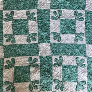 May include: A quilted textile featuring a repeating grid pattern of solid white squares and green squares with a small white floral print. The white squares are decorated with green floral-shaped appliqués and detailed diagonal stitching.
