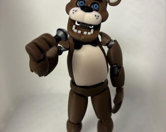 Op de afbeelding: Een bruine berenfiguur met blauwe ogen, een zwarte hoge hoed en een zwarte vlinderdas. De beer heeft een witte snuit, een witte borstvlek en vierkante tanden. Het figuur houdt een kleine grijze en zwarte microfoon in de rechterhand, met zichtbare gewrichten bij de schouders en ellebogen.