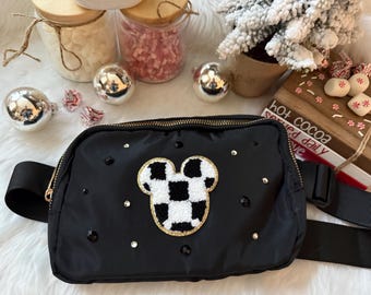 May include: Black fanny pack with a gold zipper and a Mickey Mouse head design in black and white checkered pattern. The bag is embellished with black and clear rhinestones. The adjustable strap is black.