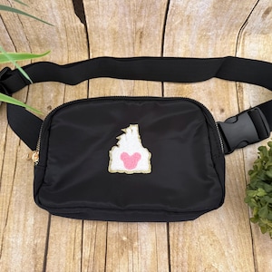 May include: Black fanny pack with a gold zipper and adjustable black strap. The pack features a white and pink patch with a gold border. The patch has a castle design with a pink heart.