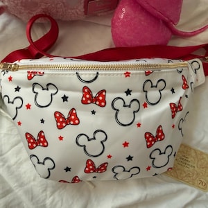K&ouml;nnte beinhalten: Wei&szlig;e Bauchtasche mit rotem Riemen und goldenem Rei&szlig;verschluss. Die Tasche ist mit schwarzen Mickey Mouse-K&ouml;pfen, roten Schleifen und roten Sternen gemustert. Ein Etikett mit der Aufschrift "Grand Californian" ist angebracht.