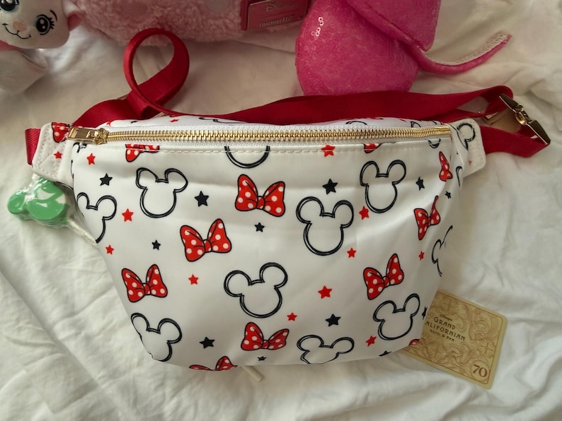 K&ouml;nnte beinhalten: Wei&szlig;e Bauchtasche mit rotem Riemen und goldenem Rei&szlig;verschluss. Die Tasche ist mit schwarzen Mickey Mouse-K&ouml;pfen, roten Schleifen und roten Sternen gemustert. Ein Etikett mit der Aufschrift "Grand Californian" ist angebracht.