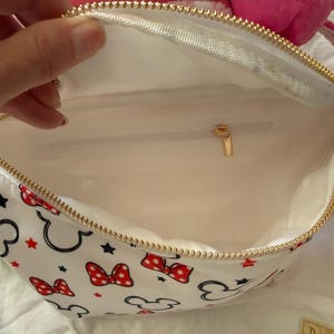 K&ouml;nnte beinhalten: Wei&szlig;e Bauchtasche mit goldenem Rei&szlig;verschluss, mit einem Muster aus schwarzen Mickey Mouse-Silhouetten, roten Schleifen und roten Sternen. Der Innenraum ist wei&szlig; mit einer kleinen Tasche und einem goldenen Rei&szlig;verschlussanh&auml;nger. Die Tasche ist offen.