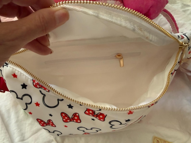 K&ouml;nnte beinhalten: Wei&szlig;e Bauchtasche mit goldenem Rei&szlig;verschluss, mit einem Muster aus schwarzen Mickey Mouse-Silhouetten, roten Schleifen und roten Sternen. Der Innenraum ist wei&szlig; mit einer kleinen Tasche und einem goldenen Rei&szlig;verschlussanh&auml;nger. Die Tasche ist offen.