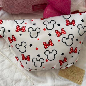 K&ouml;nnte beinhalten: Wei&szlig;e Bauchtasche mit einem Muster aus schwarzen Mickey Mouse-K&ouml;pfen, roten Schleifen mit Punkten und roten und schwarzen Sternen. Die Tasche hat einen roten Riemen und einen roten Rei&szlig;verschluss. Ein Disney-Accessoire.
