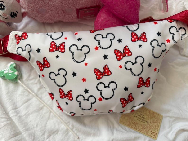 K&ouml;nnte beinhalten: Wei&szlig;e Bauchtasche mit einem Muster aus schwarzen Mickey Mouse-K&ouml;pfen, roten Schleifen mit Punkten und roten und schwarzen Sternen. Die Tasche hat einen roten Riemen und einen roten Rei&szlig;verschluss. Ein Disney-Accessoire.