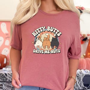 Può includere: T-shirt color malva con la scritta "Kitty Butts Drive Me Nuts" in stile retrò. Il disegno presenta tre sederi di gatti bianchi, arancioni e neri, su un arco arcobaleno con cuori e stelle.