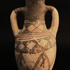 Puede incluir: Un jarrón de cerámica antigua de color beige con dos asas. Presenta patrones geométricos en negro, rojo y blanco. La parte superior y el interior son de color marrón oscuro. El jarrón está sobre un fondo negro.