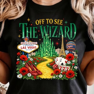 May include: Black t-shirt with a colorful graphic featuring the text "OFF TO SEE THE WIZARD." The design includes the Emerald City, a yellow brick road, red shoes, and other iconic imagery. The shirt also has the text "Welcome to Fabulous Las Vegas Nevada."
