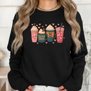 Puede incluir: Sudadera negra con un dise&ntilde;o de cuatro bebidas ilustradas con crema batida, corazones y pajitas. Una taza tiene el texto "DUTCH BROS". El dise&ntilde;o es en rosa, turquesa y marr&oacute;n. Peque&ntilde;os corazones rosas est&aacute;n dispersos.