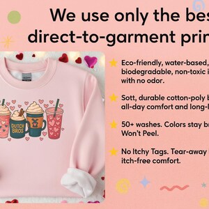 Puede incluir: Una sudadera rosa claro con un gr&aacute;fico de cuatro bebidas de caf&eacute; con crema batida y corazones. El texto "DUTCH BROS" est&aacute; en una de las tazas. La imagen tambi&eacute;n incluye texto sobre la impresi&oacute;n directa a la prenda.