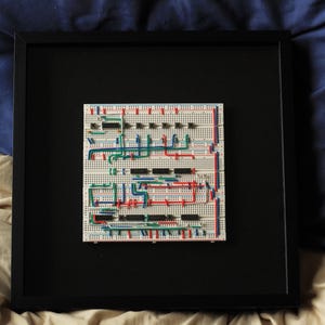 May include: A framed circuit board art piece. The circuit board is white with red, green, and blue wires connecting black integrated circuits. The frame is black and square.