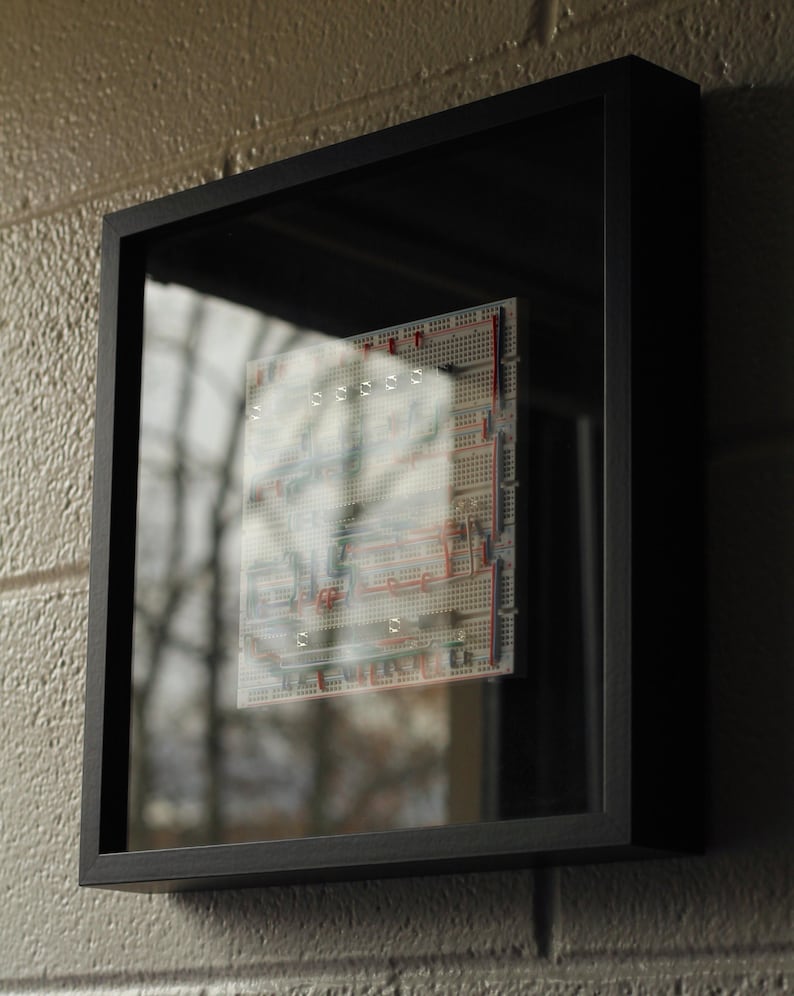 Op de afbeelding: Een zwart omkaderde printplaat met rode, blauwe en witte componenten. Het frame is gemonteerd op een grijze bakstenen muur. Het glas reflecteert een vage buitensc&egrave;ne. Het ontwerp is modern en technisch ge&iuml;nspireerd.