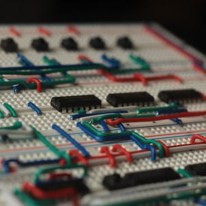 Op de afbeelding: Close-up van een wit breadboard met diverse elektronische componenten en gekleurde draden. De draden zijn rood, groen en blauw en verbinden zwarte ge&iuml;ntegreerde circuits en andere kleine zwarte componenten. Het bord wordt gebruikt voor prototyping.