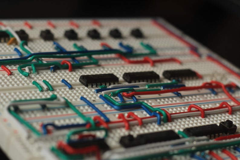 Op de afbeelding: Close-up van een wit breadboard met diverse elektronische componenten en gekleurde draden. De draden zijn rood, groen en blauw en verbinden zwarte ge&iuml;ntegreerde circuits en andere kleine zwarte componenten. Het bord wordt gebruikt voor prototyping.