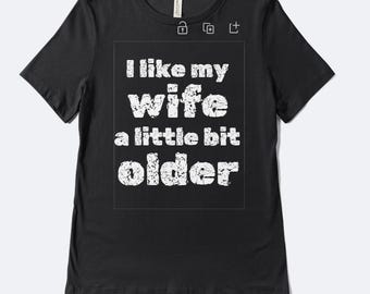 Könnte beinhalten: Schwarzes T-Shirt mit dem Text "I like my wife a little bit older" in verblichenen weißen Buchstaben. Das Shirt hat einen Rundhalsausschnitt und kurze Ärmel. Der Text ist mittig auf der Vorderseite des Shirts platziert.