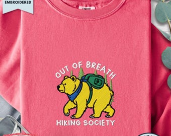 May include: A watermelon pink crewneck sweatshirt featuring an embroidered design of a yellow bear wearing a blue bandana and a green backpack. The text "OUT OF BREATH HIKING SOCIETY" is embroidered in white above and below the bear, with green pine trees in the background.