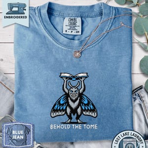 Op de afbeelding: Een blauw t-shirt met korte mouwen voorzien van een geborduurde afbeelding van een grijs en blauw motachtig wezen dat een open boek boven zijn hoofd houdt, met de tekst BEHOLD THE TOME in het wit onder het wezen op de borst.