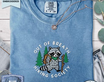 May include: A blue short-sleeved t-shirt featuring an embroidered graphic of a raccoon wearing a backpack and holding a wooden hiking stick, surrounded by green pine trees and the text "OUT OF BREATH HIKING SOCIETY" in white lettering.