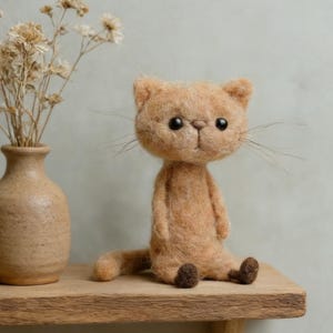 Könnte beinhalten: Eine handgefertigte, hellbraune Filzkatzenpuppe mit dunkelbraunen Augen und Schnurrhaaren sitzt auf einem Holzregal. Links von der Katze befindet sich eine kleine, beige Vase mit getrockneten Blumen. Die Katze hat eine sitzende Pose mit ausgestreckten Beinen.