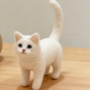 May include: A handcrafted white cat figurine with blue eyes and a raised tail. The cat is made of a soft, textured material and stands on four legs. A small, brown vase with dried flowers is in the background.