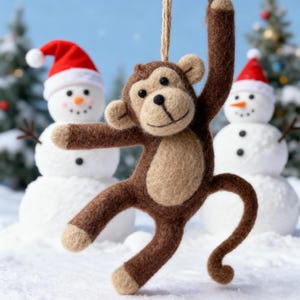 May include: A brown felt monkey ornament with tan accents, hanging from a natural fiber string. The monkey is posed with one arm raised. Two snowmen wearing red Santa hats are in the background, creating a festive winter scene.