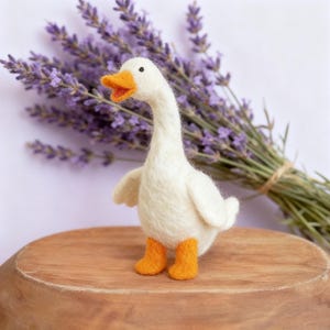 Op de afbeelding: Een handgemaakt wit ganzenbeeldje met een oranje snavel en poten. De gans is gemaakt van zacht materiaal en staat op een houten oppervlak, met een bosje paarse lavendel op de achtergrond.
